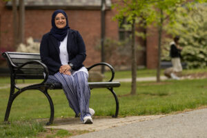 A woman sits on a bench alongside a path outside in front of a brick building. There are grass and trees in the background as well.