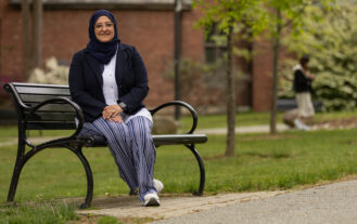 A woman sits on a bench alongside a path outside in front of a brick building. There are grass and trees in the background as well.