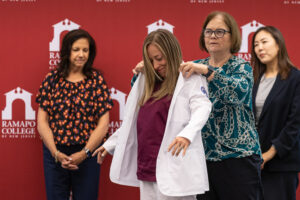 A woman stands while another woman puts a white nursning coat on her. Two other women look on. They are all in front a a maroon wall with Ramapo College logos.