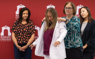 A woman stands while another woman puts a white nursning coat on her. Two other women look on. They are all in front a a maroon wall with Ramapo College logos.