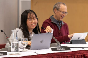 A woman sits at a table with a laptop and microphone in frotn of her. She is talking into the microphone. A man sits to her right.