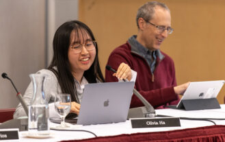 A woman sits at a table with a laptop and microphone in frotn of her. She is talking into the microphone. A man sits to her right.