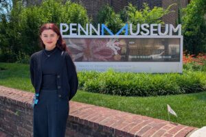 A woman stands outside in front of a sign that reads "Penn Museum".