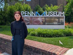 A woman stands outside in front of a sign that reads "Penn Museum".