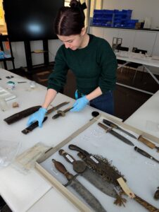 A woman wearing plastic gloves stands at a table and looks down at historical objects as she touches them.