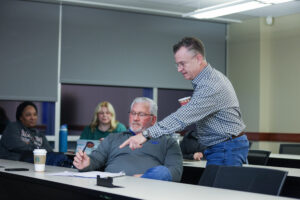An older man sits at a table in a classroom as another man points down to a paper on the table. Two woman sit at tables in the background.