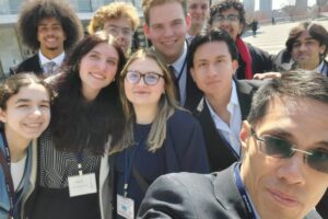 A selfie of about ten people dressed in suits. They are outside with tall buildings behind them.