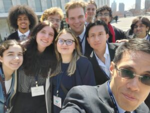 A selfie of about ten people dressed in suits. They are outside with tall buildings behind them.