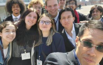 A selfie of about ten people dressed in suits. They are outside with tall buildings behind them.
