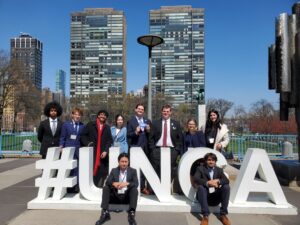 About ten people dressed in business suits pose outside near large white letters. Tall buildings are behind them.