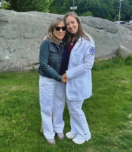 Two woman stand together and hold hands. The woman on the right has on a white nursing coat.