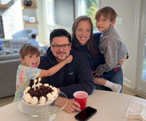 A family, including a mom, dad and two boys pose in front of a birthday cake. They are at a kitchen table.