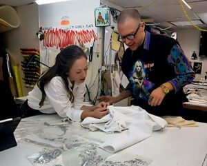 A woman works on a garment on a table. A man stands beside her.