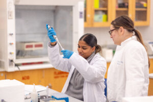 Two students in white lab coats are working in a science lab. One is wearing blue latex gloves and piping liquid into a beaker while the student to the right watches.