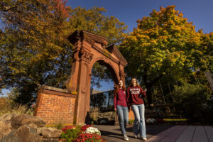 Two students in jeans and maroon tops are walking towards the camera. The Havemeyer Arch is behind them with colorful fall foliage on trees and a clear blue sky.