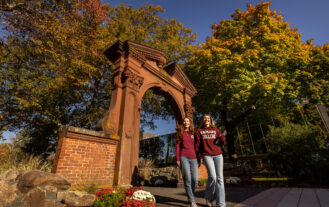 Two students in jeans and maroon tops are walking towards the camera. The Havemeyer Arch is behind them with colorful fall foliage on trees and a clear blue sky.