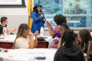 A woman stands at the front of a room holding a microphone. In front of her are college students seated at round tables.
