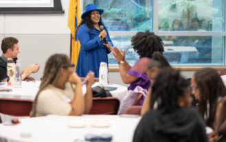 A woman stands at the front of a room holding a microphone. In front of her are college students seated at round tables.