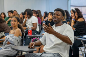 A young man sits in a classroom, speaking and gesturing with his hands. Other students are seated in desks around him.