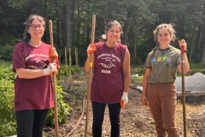 Three young women stand outside holding rakes. They are smiling.