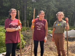 Three young women stand outside holding rakes. They are smiling.