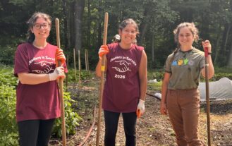 Three young women stand outside holding rakes. They are smiling.