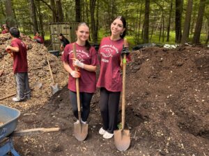 Two young women hold shovels in front of a pile of mulch. They are wearing Ramapo College shirts and smiling.