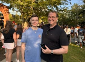 A father and college-aged son stand together outside and smile.