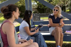 A young woman with headphones and a microphone sits down and points the microphone toward another woman on a bench. Another young woman is beside her, also looking at the interview subject. They are all outside.