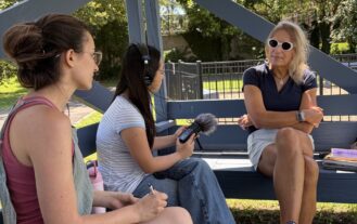 A young woman with headphones and a microphone sits down and points the microphone toward another woman on a bench. Another young woman is beside her, also looking at the interview subject. They are all outside.