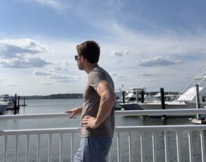 A man stands leans against a white fence at a marina. He is looking at the water and boats, and his side is toward the camera.