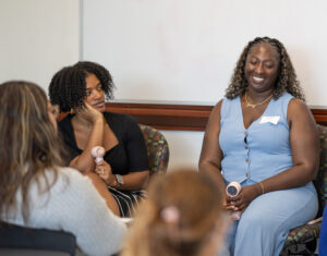 Two women sit on chairs in the front of a room. One smiles while the other looks at her. They are in front of two women.