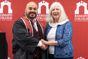 A man in a graduation gown and stole shakes hands with a woman and smiles. They are in front of a Ramapo College of New Jersey backdrop.