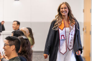 A young woman with a graduation gown and a nursing stole beams a big smile as she walks inside a room.