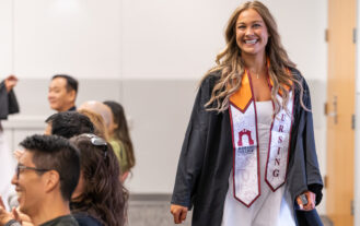 A young woman with a graduation gown and a nursing stole beams a big smile as she walks inside a room.