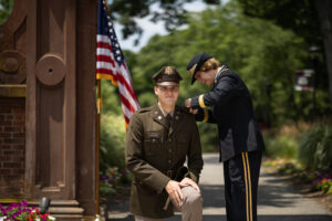 A young man in a military uniform kneels down outside in front of an American flag, as a woman in a military uniform pins a pin onto his shoulder.