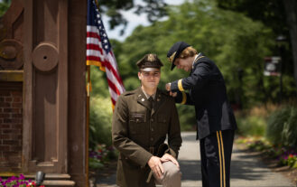 A young man in a military uniform kneels down outside in front of an American flag, as a woman in a military uniform pins a pin onto his shoulder.