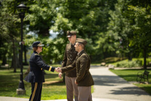 A woman in a military uniform shakes the hand of a young man in a military uniform as another young man in uniform looks on. They are all outside in front of green trees.