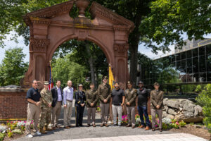 A group of people, some in military uniforms, stand shoulder to shoulder in front of a large arch outside.