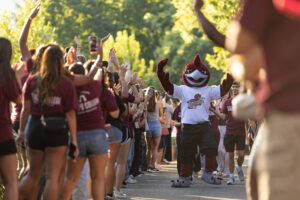 A large Rpadrunner mascot walks with its hands up. It is surrounded by college students in maroon shirts.