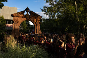 A group of young people in maroon shirts line up outside in front of a large arch. They are smiling and clapping.