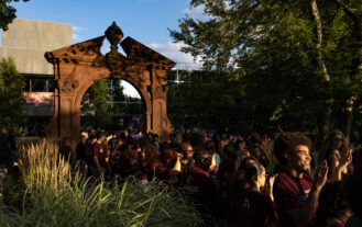 A group of young people in maroon shirts line up outside in front of a large arch. They are smiling and clapping.
