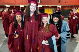 A group of three women in maroon graduation caps and gowns stand together, next to an older woman in a light blue graduation gown and black cap. They are all smiling.
