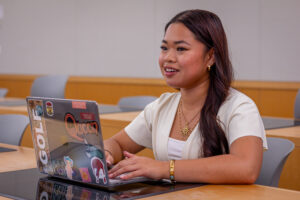 A young women sits in a classroom. Her laptop is open on the desk and she is looking ahead.