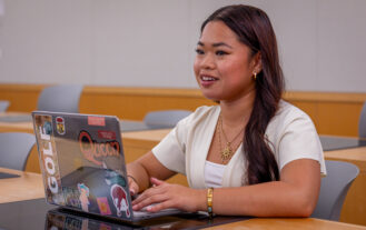 A young women sits in a classroom. Her laptop is open on the desk and she is looking ahead.