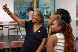 A woman holds out a phone and takes a selfie with three college students.