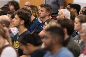 A crowd of people sit in chairs. A profile shot of a young man is in focus.