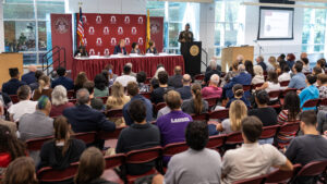 A man in a military uniform stands at a podium and speaks to a crowd. He is next to a panel of people, with a bacground behind them that states Ramapo College of New Jersey.