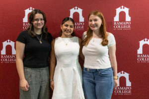 Three young women stand side by side in front of a maroon backdrop with Ramapo College logos on it.