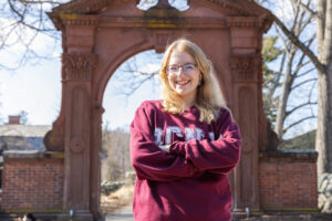 A student wearing a maroon RCNJ sweatshirt is smiling with her arms crossed while standing in front of the Havemeyer Arch. The trees are bare and the sky is blue with streaks of clouds.
